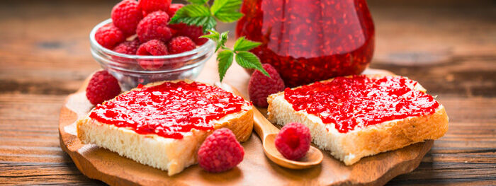 Weißbrot mit Himbeermarmelade auf Holzbrett, Himbeeren in Schale Weißbrot mit Himbeermarmelade auf Holzbrett, Himbeeren in Schale